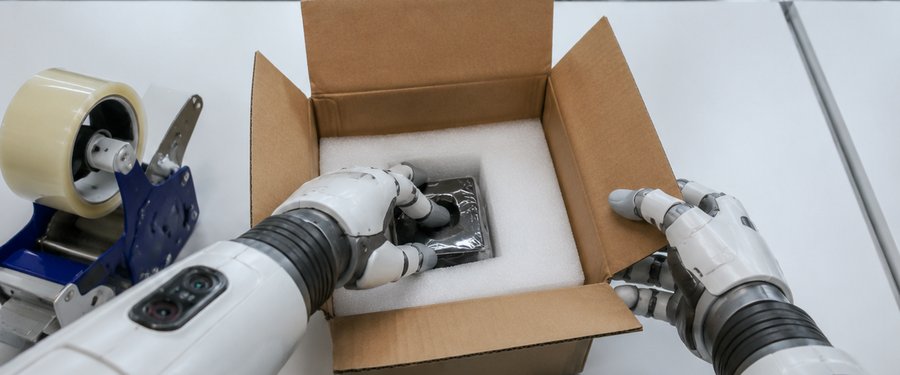 Two robot arms placing a part into a foam-lined carton on a packing bench